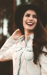 A woman wearing a hat and jeans smiles warmly, exuding a cheerful and relaxed demeanor.