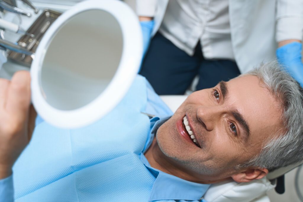 A man smiles while looking into a hand mirror at a dental office.