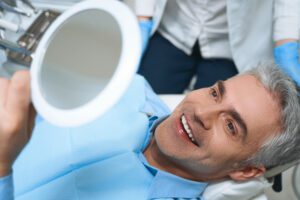 A man smiles while looking into a hand mirror at a dental office.