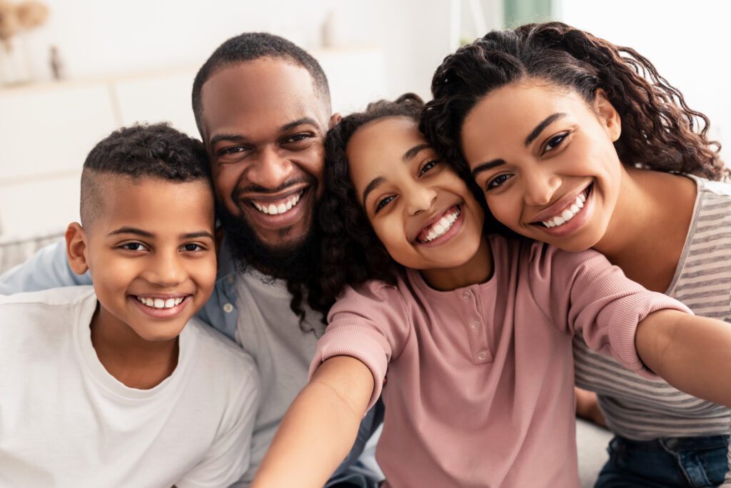 A smiling group of four people, including two adults and two children, standing close together in an indoor setting.