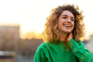 A young woman with curly hair and a green shirt smiling happily with cosmetically enhanced teeth.
