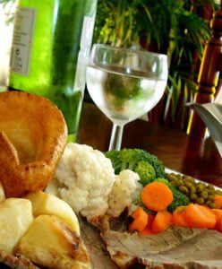 Plate of roasted meat and vegetables with a Yorkshire pudding, alongside a glass of liquid and a green bottle on the table.
